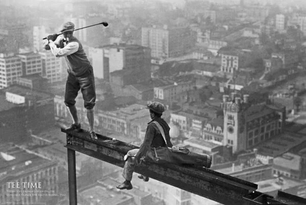 A man is swinging a golf club on a steel beam high above a cityscape, wearing vintage clothing. Another man sits nearby with a golf bag, watching. Buildings and a clock tower are visible in the foggy distance below.