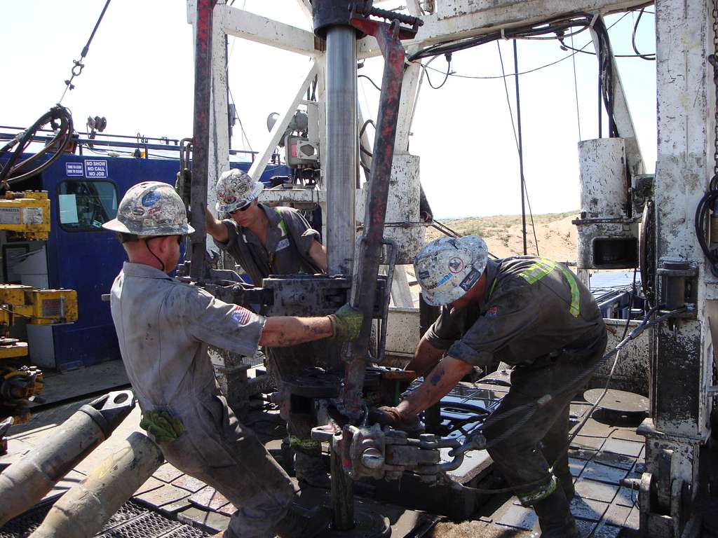 Three oilfield workers operating drilling equipment at an outdoor drilling site. They are wearing helmets and work clothes, surrounded by machinery and tools. The environment appears industrial and rugged.