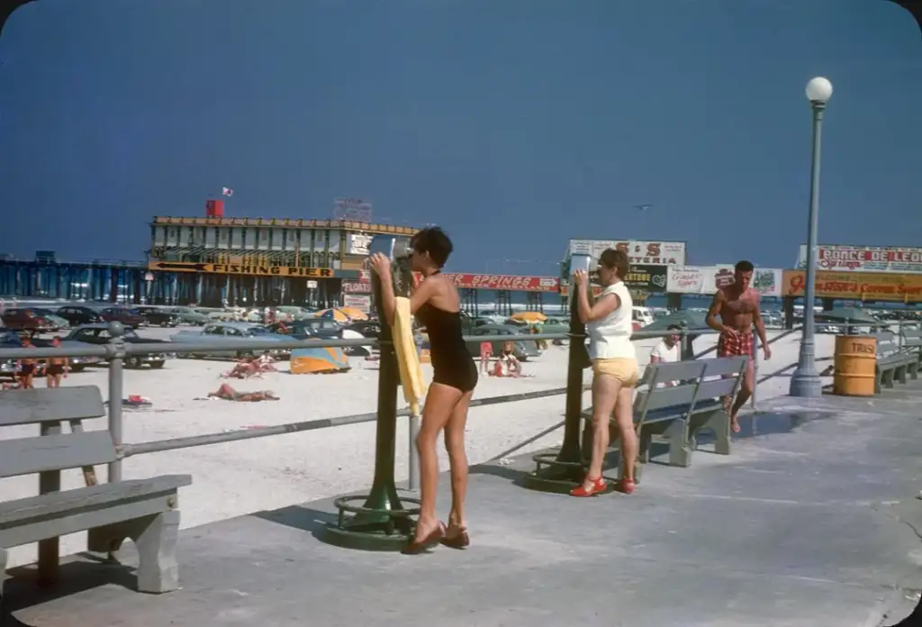 People on a boardwalk at the beach. A woman in a black swimsuit dries off, a man carries a yellow towel, and a woman in a white top looks on. There are benches, parked cars, and a pier in the background under a clear blue sky.