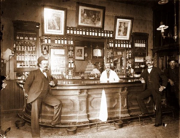 A sepia-toned image of an old-fashioned bar. Three men in suits and hats lean against the wooden bar counter, with one man behind it. Shelves filled with bottles and framed pictures adorn the wall. The atmosphere is vintage and classic.