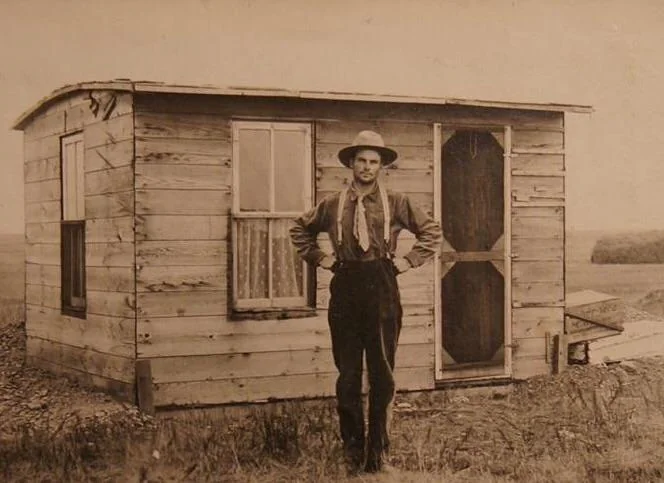 A man in a hat and suspenders stands with hands on hips in front of a small wooden shack. The building has two windows and a screen door, and it is set in an open, grassy landscape. The image is in sepia tones.