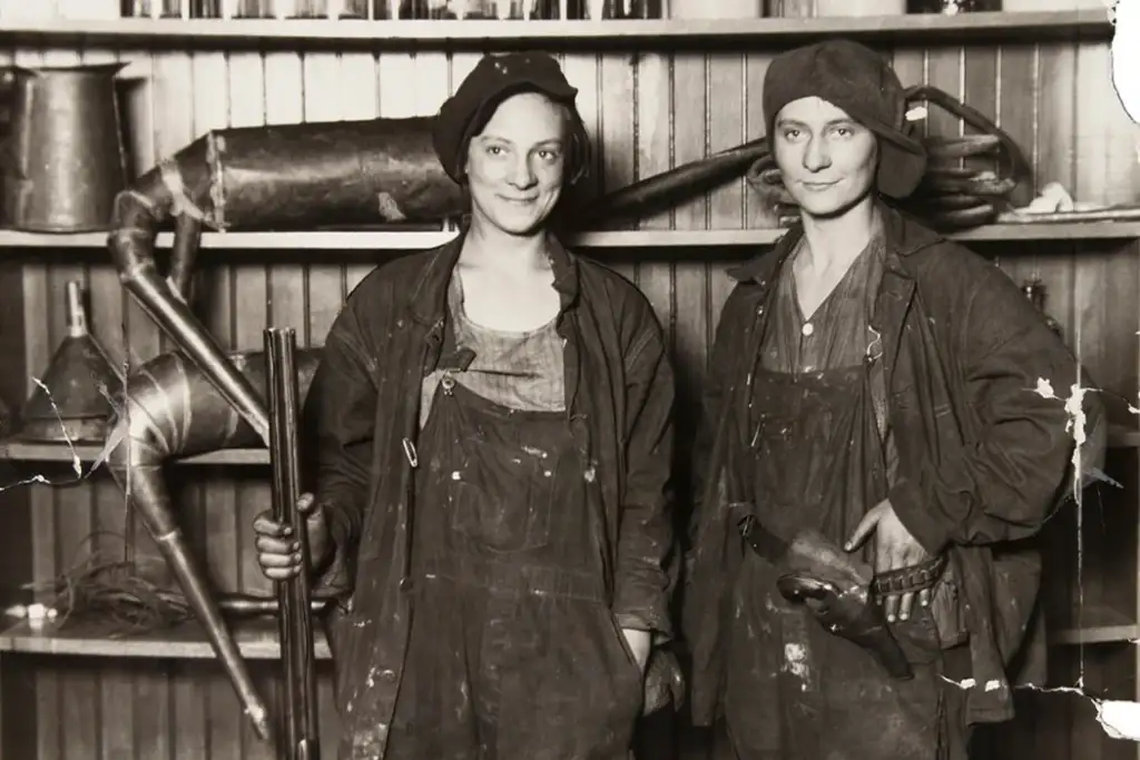 Two people in work attire stand in front of shelves with tools and equipment. Both wear dark overalls and caps. One holds a shotgun, and the other has a hand on their hip. The background consists of a wooden shelf with various objects.