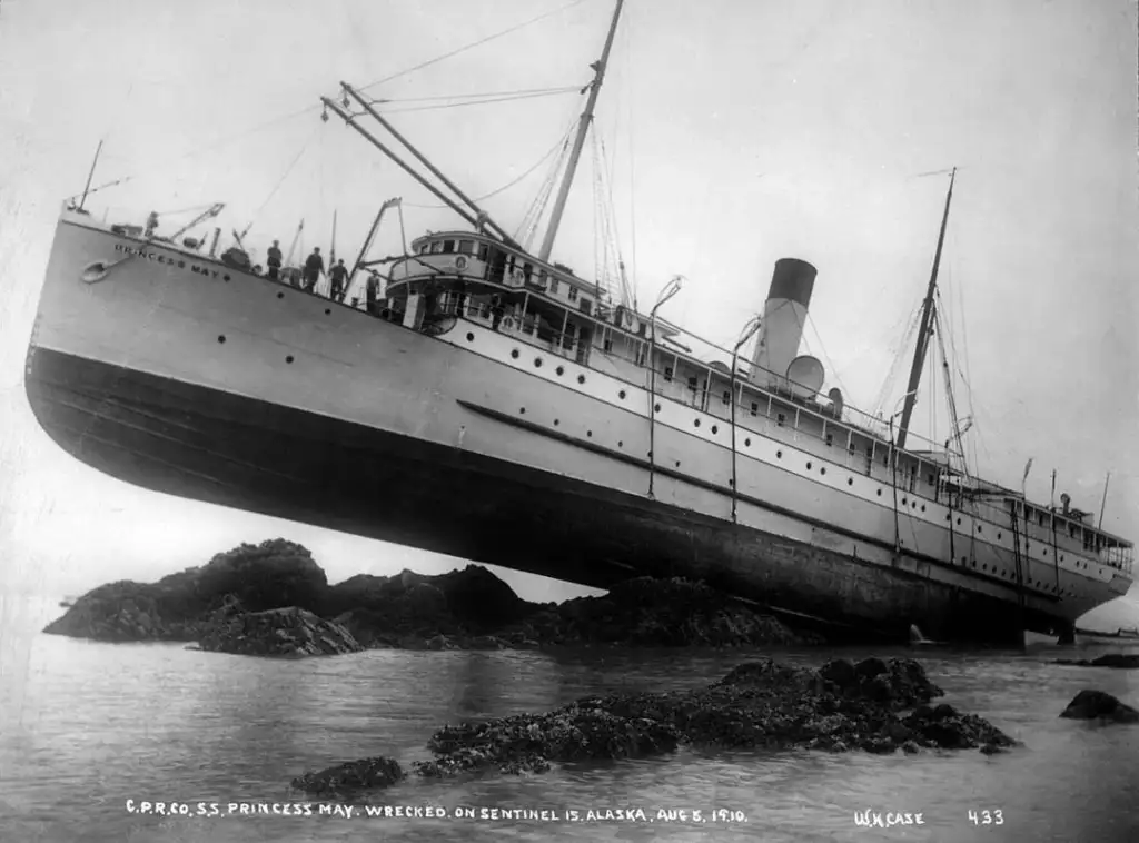 Black and white photo of the SS Princess May ship grounded on rocky terrain near Sentinel Island, Alaska, in 1910. The ship is tilted to the right with people visible on the deck.