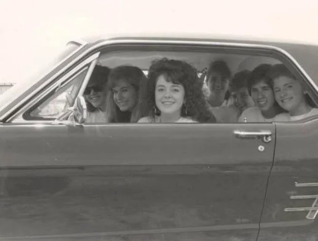 A group of seven people, four women and three men, sit inside a classic car. They are smiling and looking out the open window. The image is in black and white, suggesting a vintage or retro vibe.