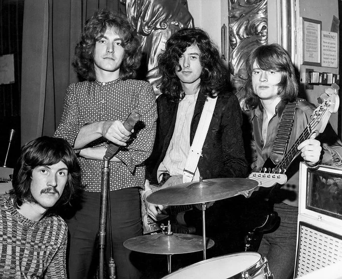 Four musicians from a rock band pose with their instruments in a vintage black and white photo. One stands next to a microphone, others hold guitars, and the drummer sits with his drum kit. Decorative wall art is partially visible in the background.