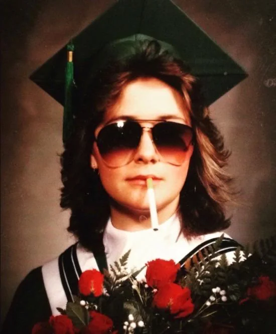 A graduate wearing a green cap and gown holds a bouquet of red roses. They have sunglasses on and a cigarette hanging from their mouth.