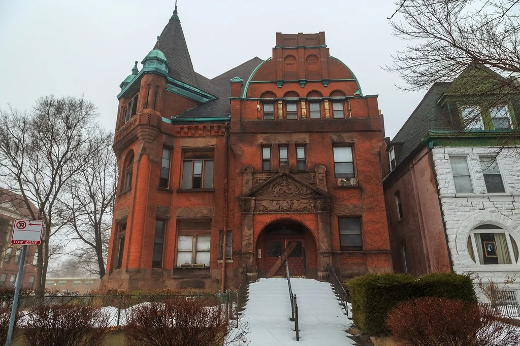 A large, historic red brick building with a pointed tower and ornate architectural details. It features a snowy front staircase leading to the entrance. Bare trees and neighboring buildings are visible on a foggy day.