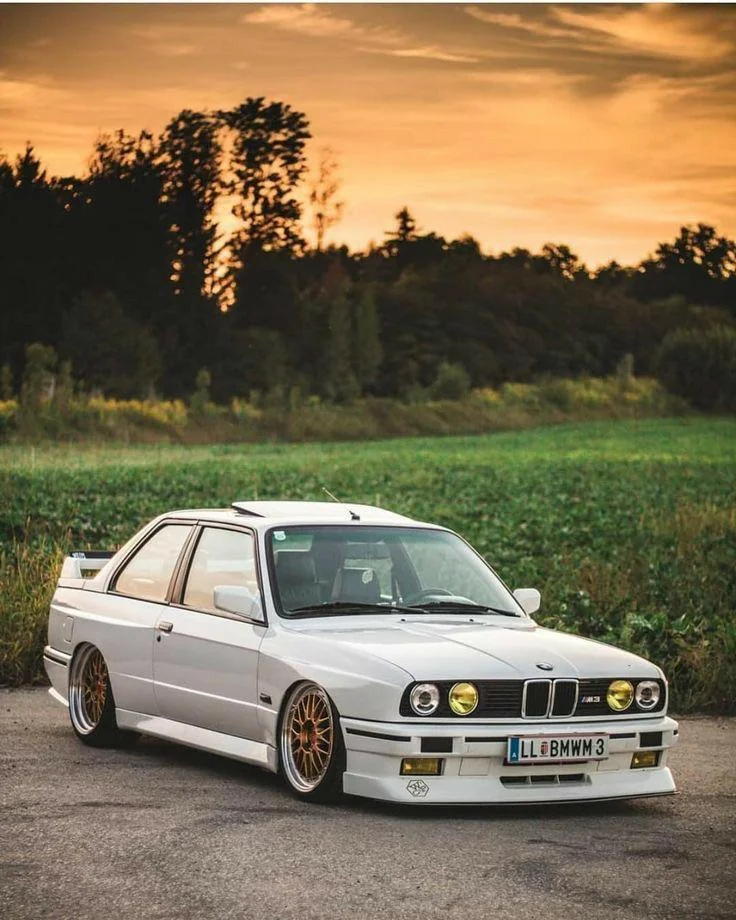 A white BMW M3 E30 is parked on a rural road at sunset. The car has gold rims and a sleek, classic design. In the background, there's a field and a line of trees silhouetted against an orange sky.