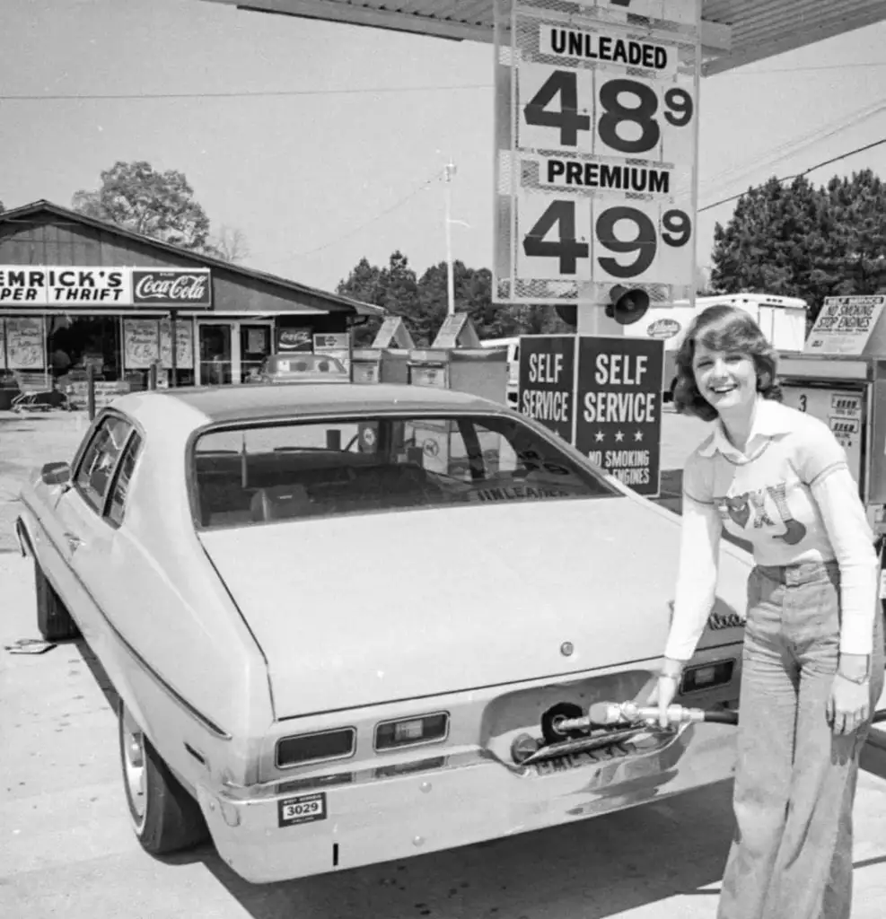 A woman stands beside a car at a gas station, smiling while refueling. Gas prices are displayed as 48.9 cents for unleaded and 49.9 cents for premium. A store with a Coca-Cola sign is visible in the background. The scene appears vintage.