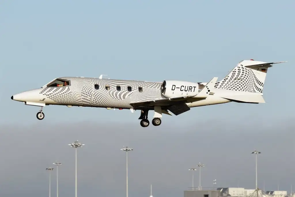 A white and black private jet with a geometric pattern is flying against a clear blue sky. The registration number "D-CURT" is visible on the side. The jet is approaching landing with the landing gear deployed.