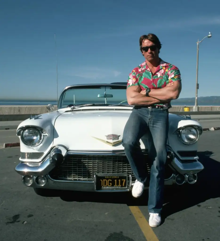 A man in a floral shirt and jeans, arms crossed, leans against a white classic convertible car parked on a street with a clear sky and a distant view of water. The car has a vintage design and a visible front license plate.