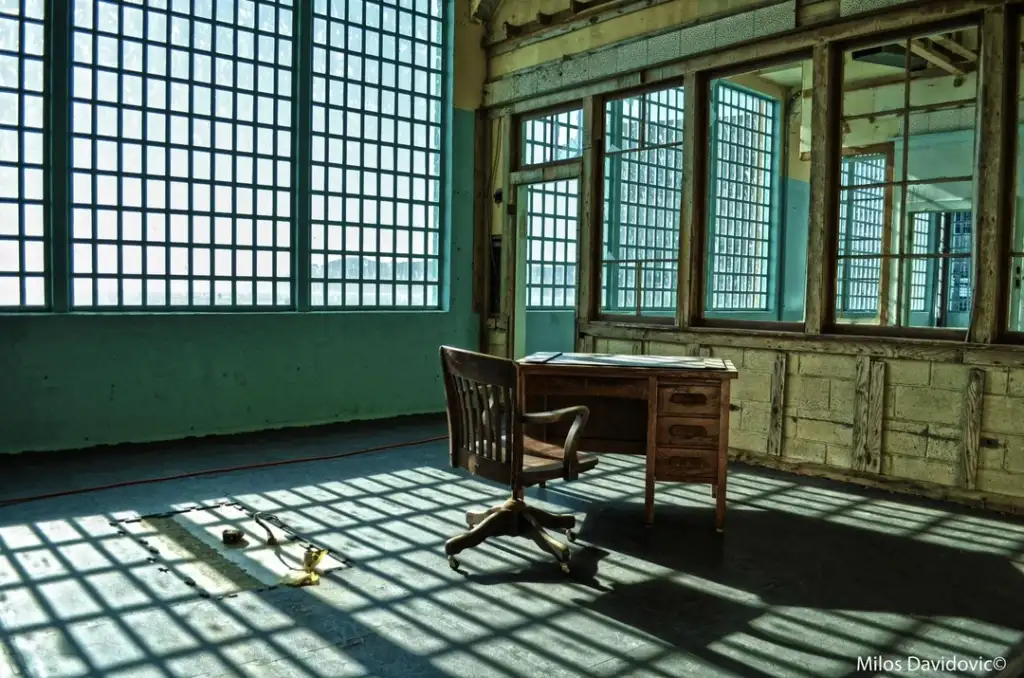 An empty room with large grid windows casting shadows on the floor. In the center, a wooden desk and chair. The walls have peeling paint, and the overall atmosphere is abandoned. Mirrors reflect the scene on one wall.