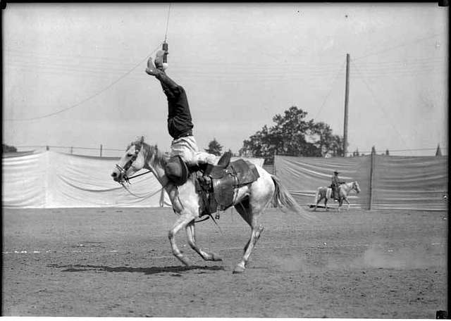 A person performs a handstand on a moving horse in an outdoor arena with a backdrop of tall wooden panels. Another person on horseback is visible in the background.