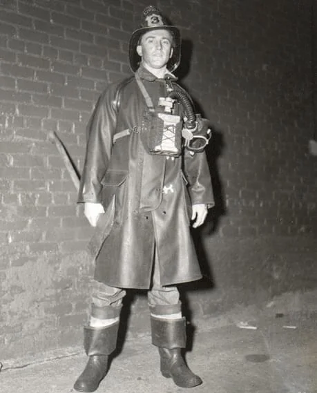 A firefighter in vintage gear stands against a brick wall. He is wearing a long coat, high boots, and a helmet with a breathing apparatus strapped to his chest. The photo is black and white, giving an old-time feel.