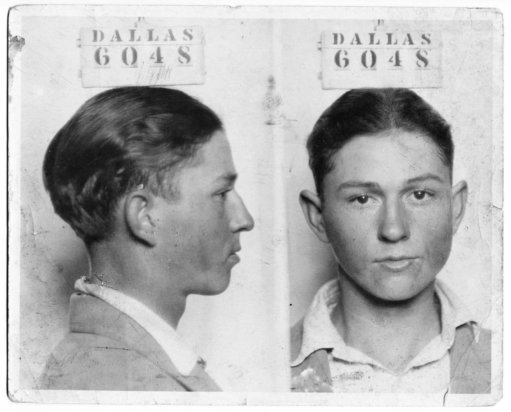 Vintage black and white mugshot of a young man with short hair. Two views are shown: a side profile on the left and a front-facing view on the right. A sign above him says "Dallas 6048." The photo has worn edges and creases.