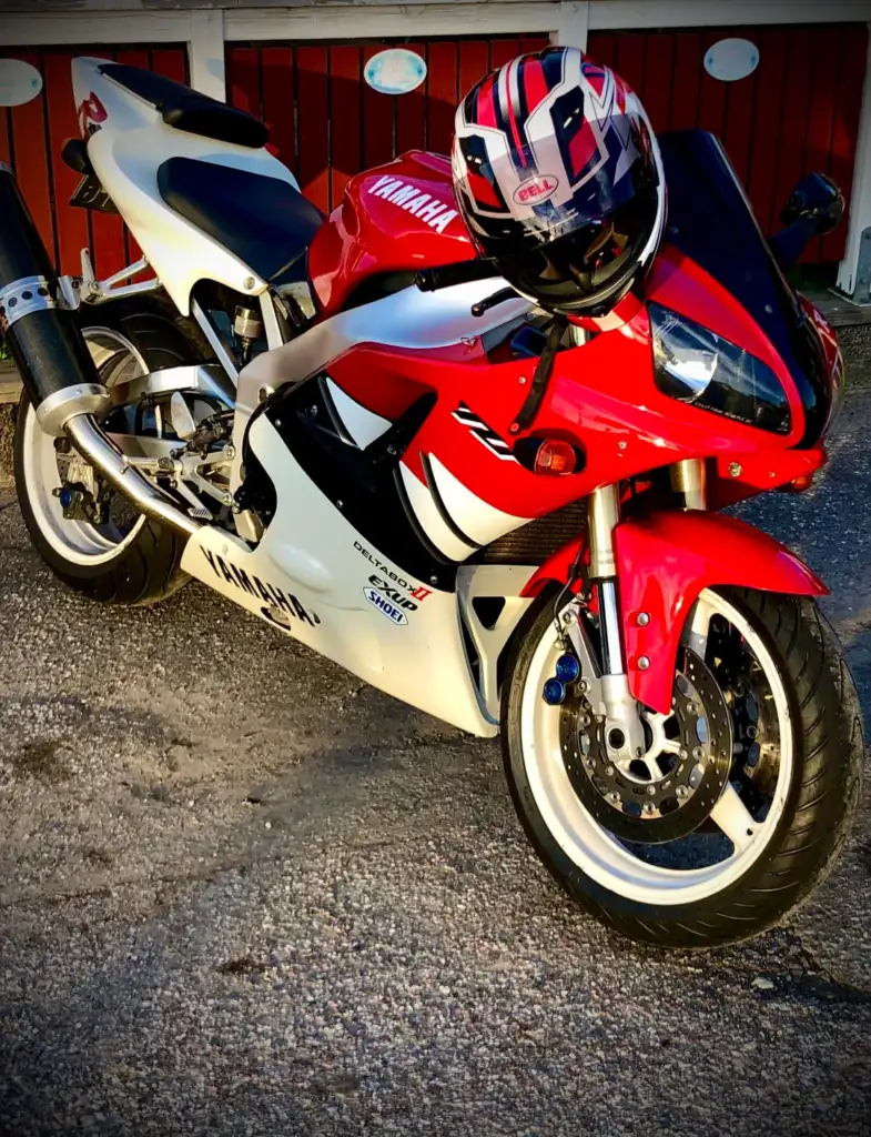 A red and white sport motorcycle is parked on a textured surface. The front fairing is visible, and a matching helmet rests on the handlebars. The bike has bold graphics and a shiny exhaust, with a red building in the background.