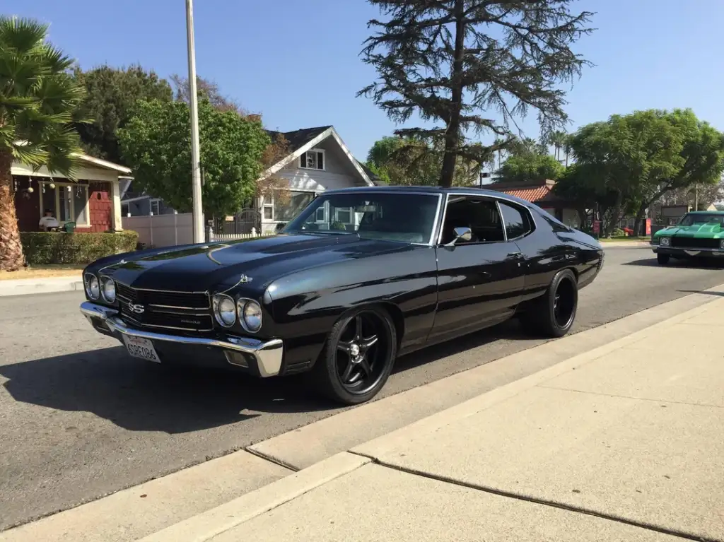 A black classic muscle car, parked on a suburban street. It has a shiny exterior with black rims and a chrome front grille. Trees and houses are in the background under a clear blue sky.