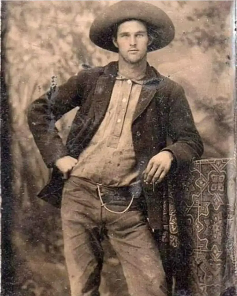 Sepia-toned photograph of a man in 19th-century attire. He is wearing a wide-brimmed hat, jacket, and bow tie, with one hand on his hip and the other resting on a patterned tablecloth. The background features a blurred nature scene.