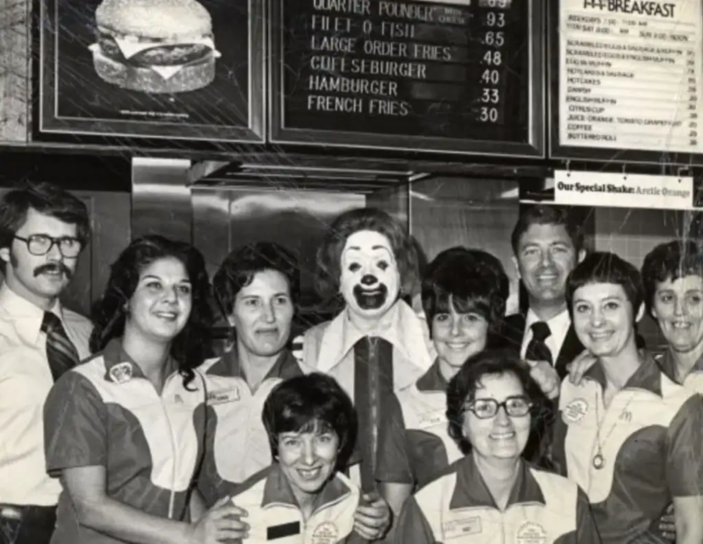 A black and white photo showing a group of smiling employees in uniform gathered in front of a fast-food counter menu. They are posing with a person dressed as a clown. The menu board lists items like hamburgers and fries.