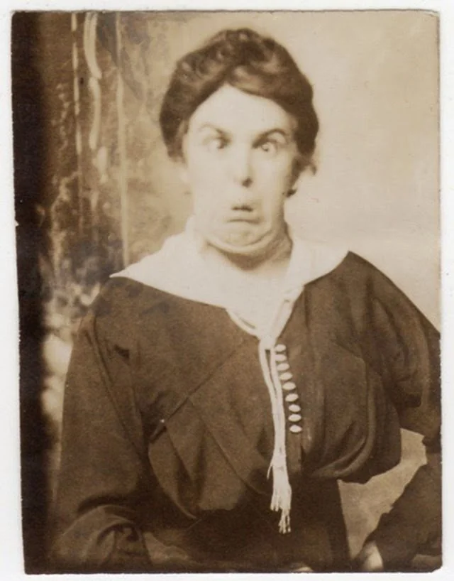 A vintage photograph of a person with short, dark hair making a comical and exaggerated facial expression, wearing a dark dress with a high collar and buttons. The background is simple and slightly blurred.