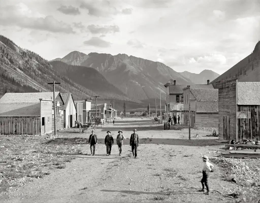 A black and white photograph depicts a group of four men walking towards the camera on a dirt road in a small town. Wooden buildings line both sides of the road, and mountains rise in the background. A child is running to the right.