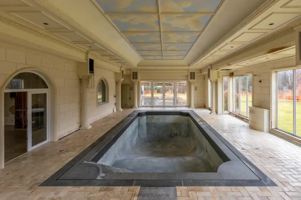 Indoor pool area with an empty pool. The walls are cream-colored with large windows on one side, allowing natural light. The ceiling is painted with clouds, adding an artistic touch. The floor is tiled in a beige pattern.