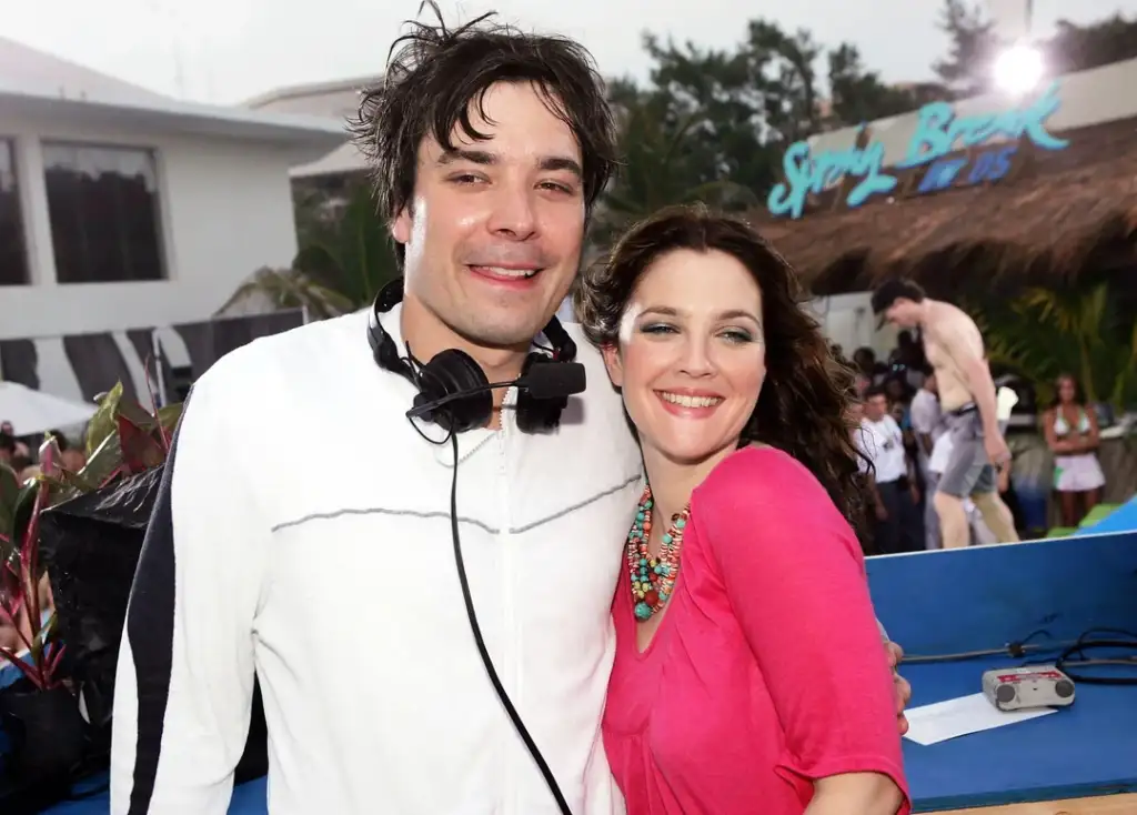 A man and woman smiling and posing together outside at an event, with people and a building in the background. The man is wearing headphones around his neck, and the woman is wearing a pink top and colorful necklace.