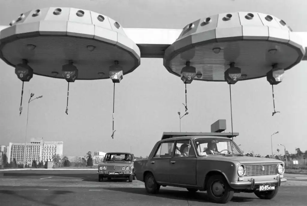 Vintage black-and-white photo of two cars passing through futuristic, UFO-like toll booths. The scene has a retro-futuristic design with circular structures and hanging elements, set against a backdrop of trees and buildings.