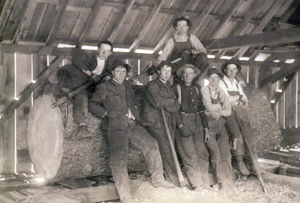 A group of seven men, dressed in early 20th-century work clothing, pose with logging tools inside a wooden structure. They lean on a large, horizontal tree trunk. Sunlight filters through the gaps in the walls and roof.