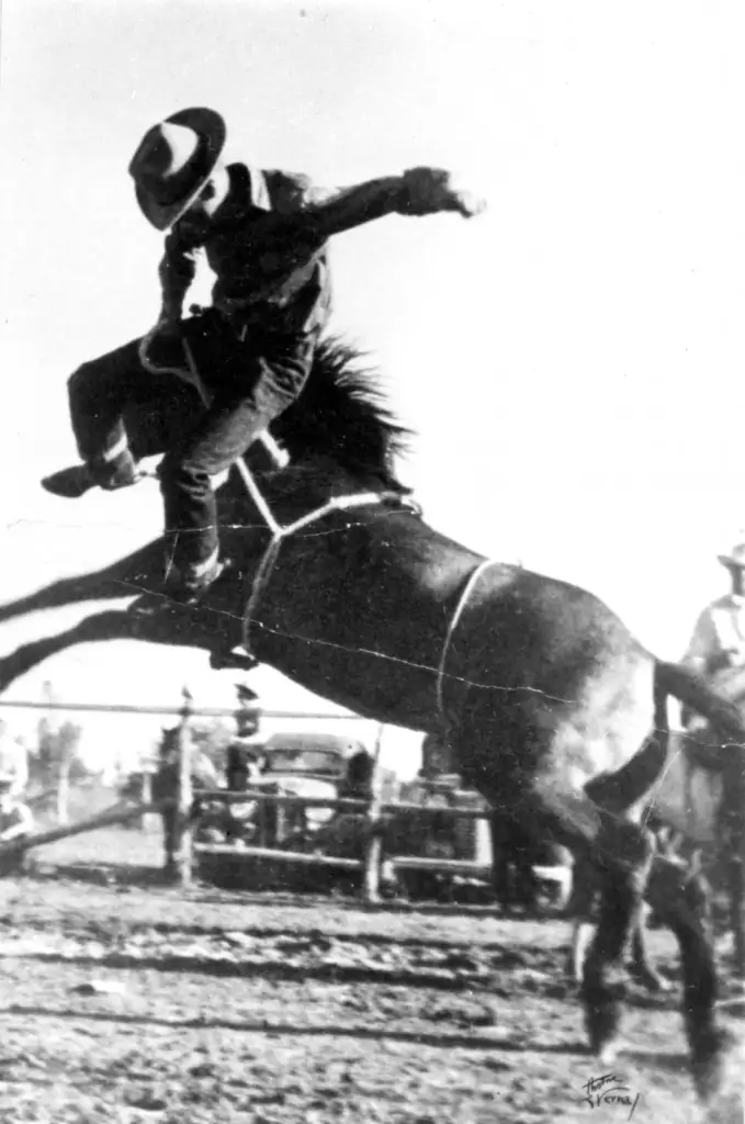 A man wearing a hat and cowboy attire rides a bucking horse in a rodeo arena. The horse is mid-jump, its mane flying. In the background, spectators watch the action from behind a fence. The image is black and white.