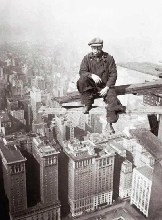 A construction worker sits on a wooden beam high above a city, with tall buildings and a hazy skyline in the background. He is wearing a cap and work clothes, exuding a sense of calm despite the height.