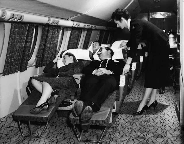 Black and white photo of four passengers sleeping comfortably on reclining seats in an aircraft, attended by a flight attendant in a uniform. The cabin interior has patterned carpeting and curtains at the windows.