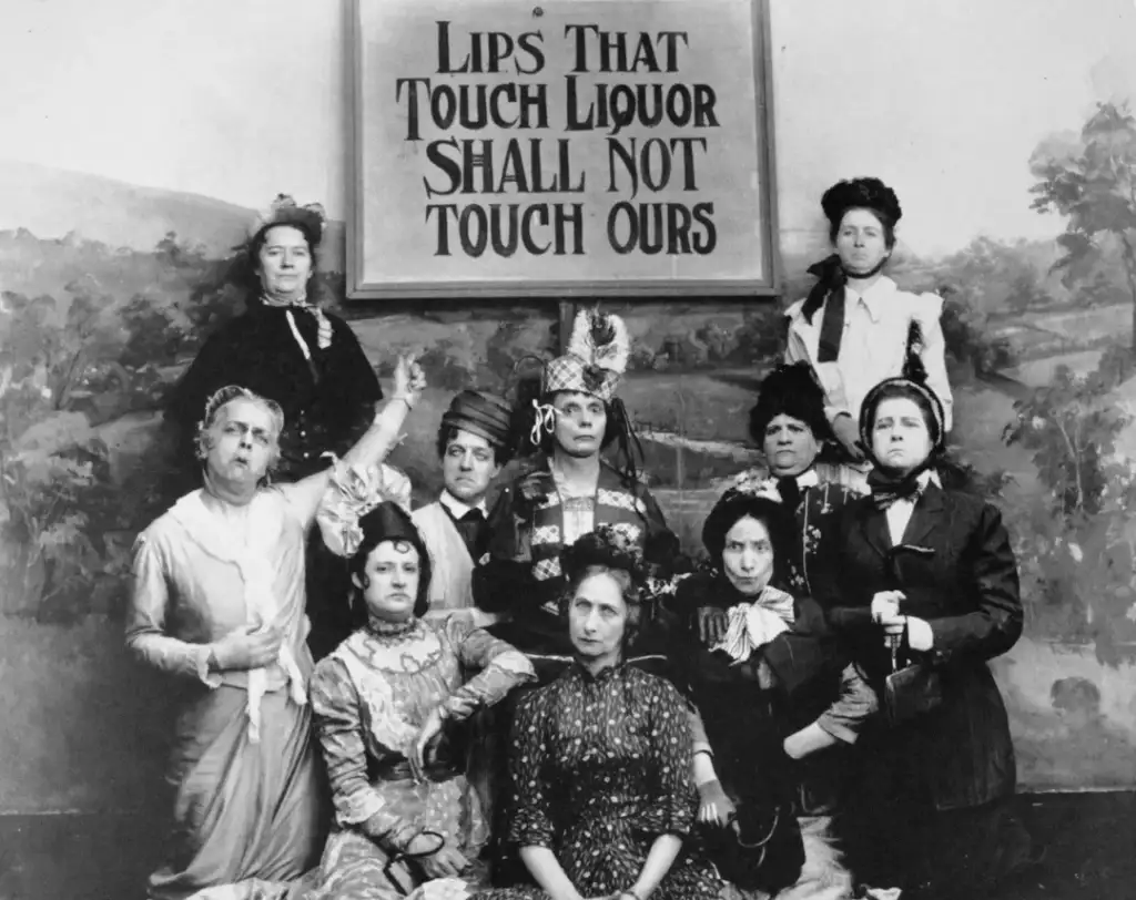 A group of ten women in period clothing pose in front of a sign that reads, "Lips That Touch Liquor Shall Not Touch Ours." Some hold stern expressions, with one pointing to the sign. The setting appears to be historical.