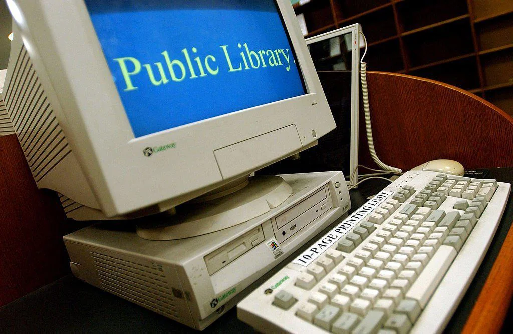 A vintage computer setup in a library displays "Public Library" on the screen. The computer tower and CRT monitor are beige, with a keyboard labeled "10.4 PAGE PRINTING GUIDE" in front. Wooden shelves are in the background.