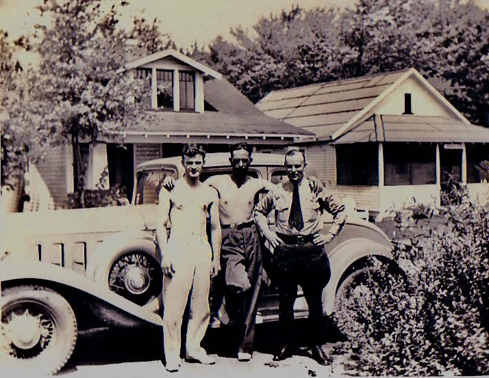 Three men stand in front of a vintage car outside a house with a porch. Two men are in shirts and pants, while the center man is shirtless. Trees are visible in the background, creating a relaxed, vintage atmosphere.