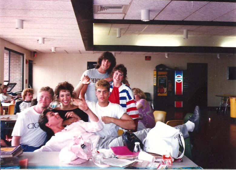 A group of people in a casual indoor setting, posing playfully around a table. One person lies across the table, while others gather around, smiling and laughing. There's a vending machine in the background. The atmosphere is friendly and relaxed.