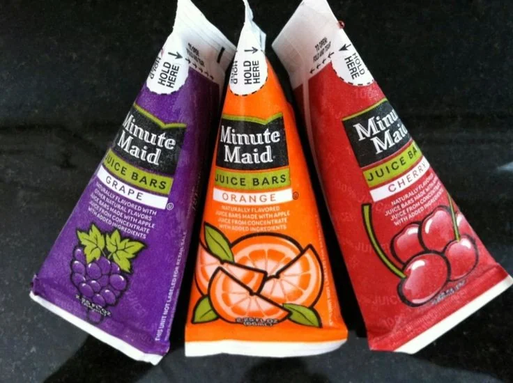 Three Minute Maid juice bar wrappers on a black surface. The flavors are grape, orange, and cherry, each with corresponding fruit illustrations. The grape wrapper is purple, the orange is orange, and the cherry is red.