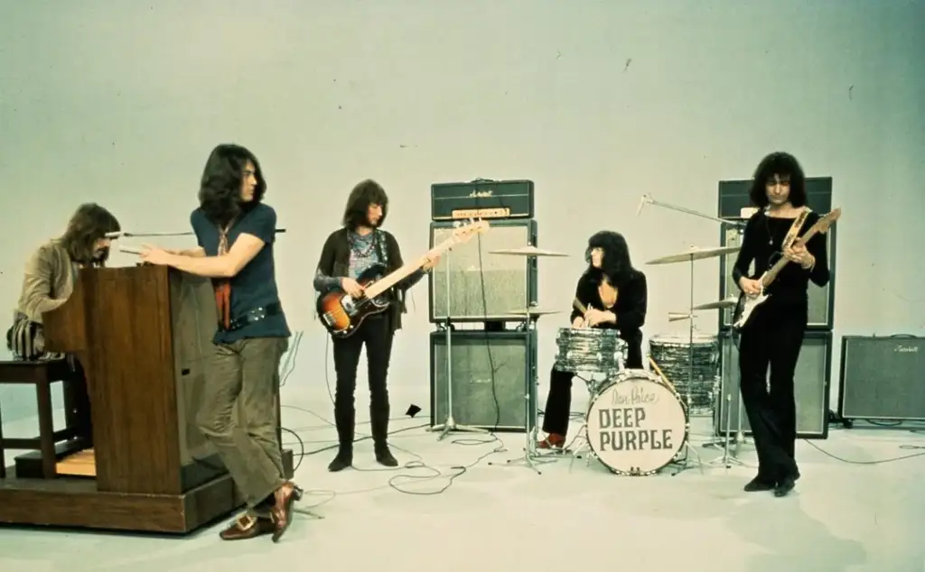 Five musicians are playing in a studio. One is on keyboards, another on bass, a third on drums, and two are playing guitars. Amplifiers are in the background, and the bass drum reads "Deep Purple." They are casually dressed.