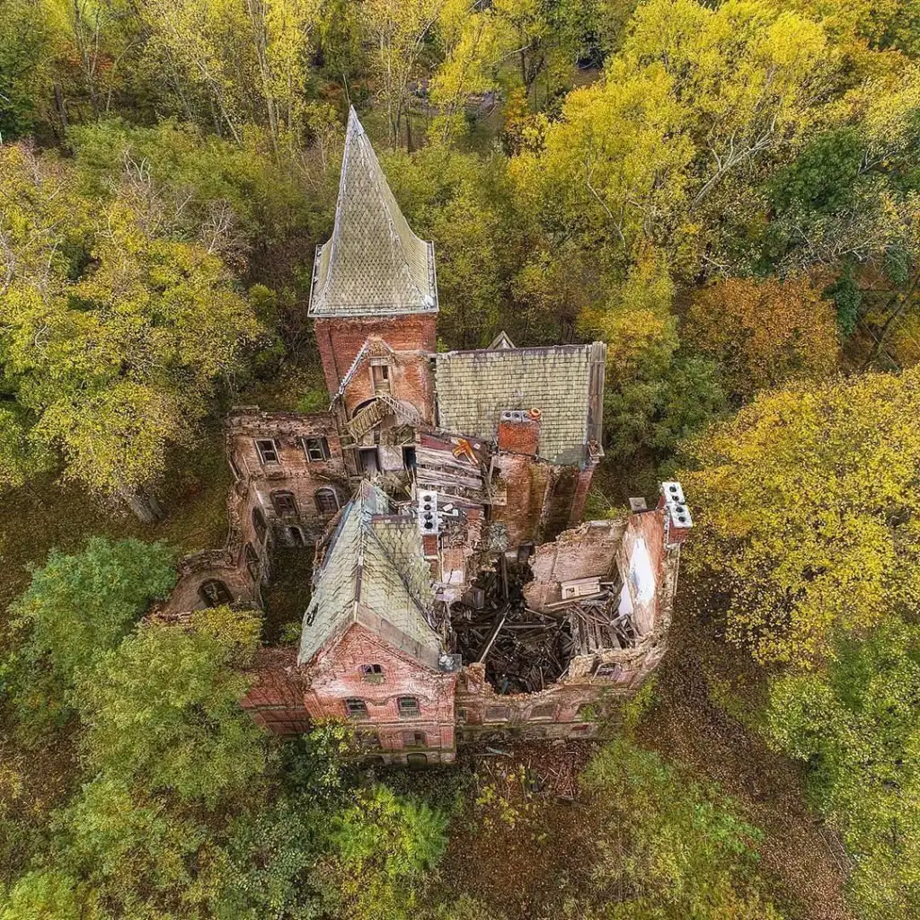 Aerial view of an abandoned, overgrown brick mansion with a tall, pointed roof and collapsed sections. Surrounded by dense trees with autumn foliage, the structure appears weathered and partly ruined, blending into the natural environment.