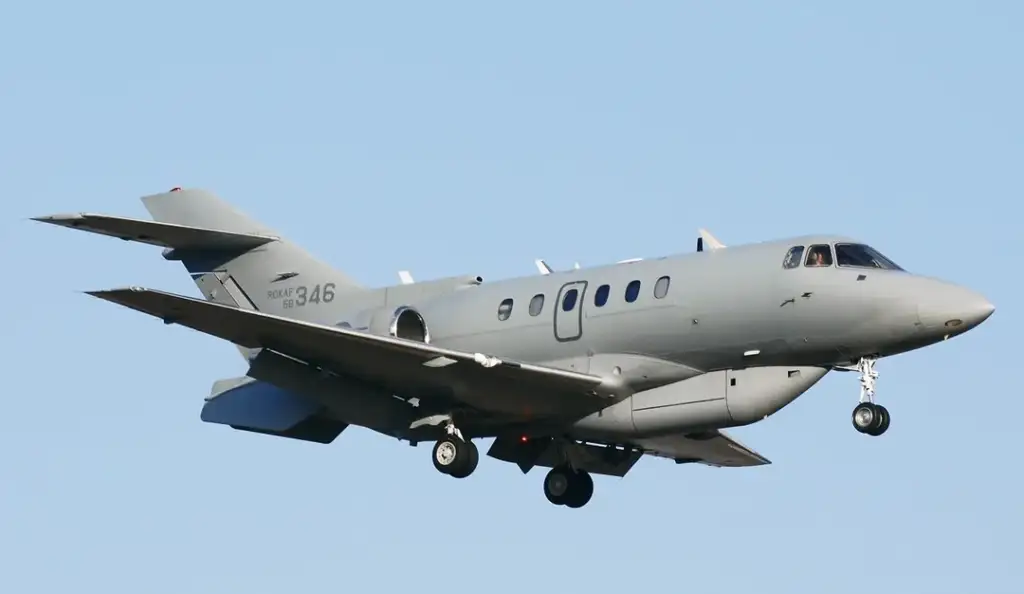 A small military jet with a gray exterior is flying in the sky. The aircraft has two engines on the wings and landing gear visible, indicating it is either taking off or preparing to land. The background shows a clear blue sky.