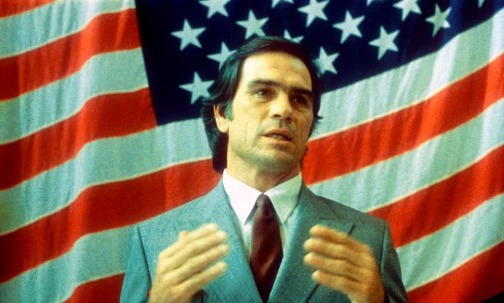 A person in a gray suit and red tie gestures with hands in front of an American flag backdrop.