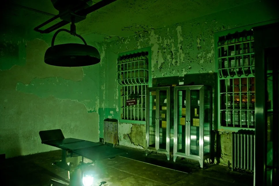 Dimly lit, eerie room with chipped green walls and barred windows. An empty medical examination chair is beneath a large overhead lamp. Two locker-like metal cabinets stand against the wall, adding to the abandoned, unsettling atmosphere.