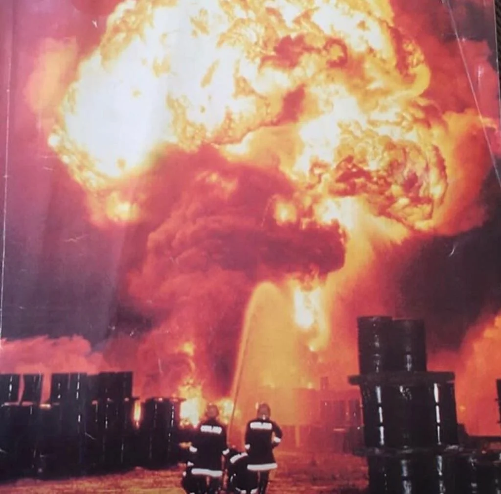 Two firefighters in protective gear battle a huge, fiery explosion with hoses. Intense flames and smoke billow, illuminating the scene. Rows of large barrels surround them, heightening the danger.