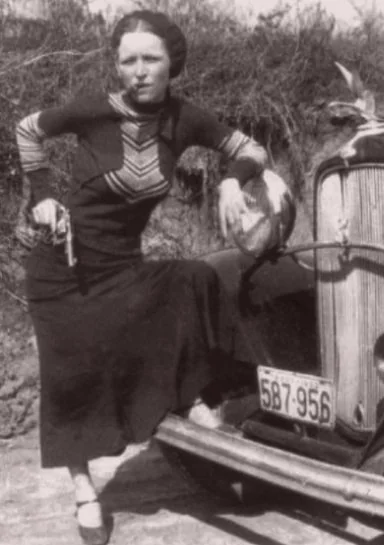 A woman in a long dark dress poses confidently with a handgun in her right hand, leaning against the front of a vintage car. She rests her left arm on the car's headlight. The background is a natural setting with bushes.