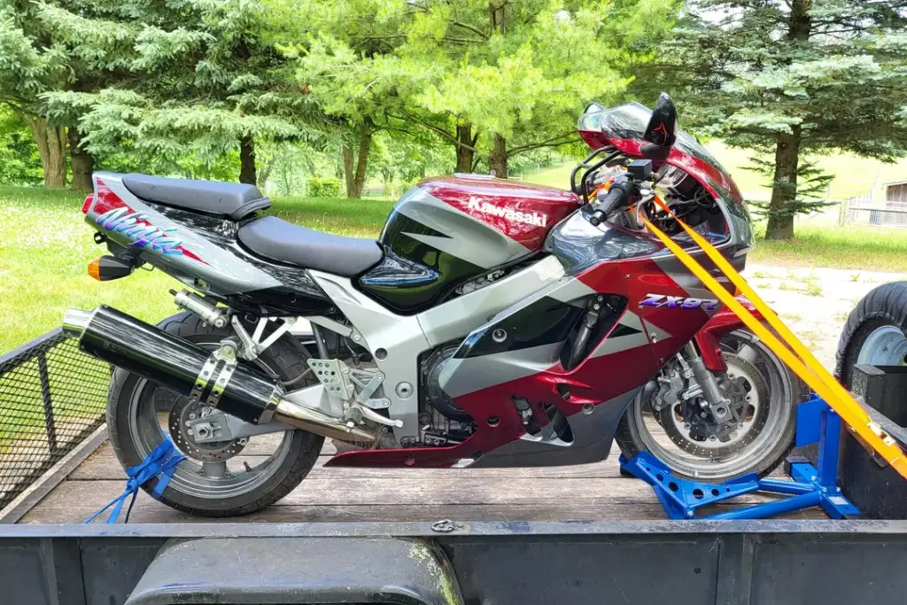 A red and silver Kawasaki ZX-9R motorcycle is secured on a trailer with orange straps. It's parked outdoors, surrounded by lush green trees. The bike has a distinctive sporty design and a large black exhaust pipe.