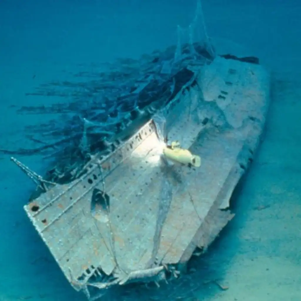 Underwater image of a shipwreck on the ocean floor, partially covered in sediment. A small submarine with a bright light is exploring the site, illuminating the ship's decaying surface and supporting structures amidst the deep blue water.