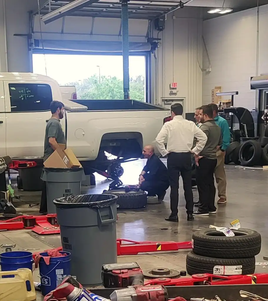 A group of five people in a garage are gathered around a vehicle with one wheel removed. Some are wearing casual attire and others are in formal clothes. The garage contains tools, tires, and equipment, with a truck parked inside.