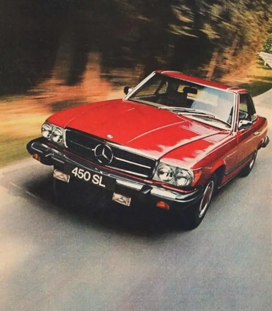 A classic red Mercedes-Benz 450 SL speeds down a scenic, tree-lined road. The car features a vintage design with a prominent Mercedes emblem on the grille, chrome accents, and sleek lines, set against a blurred green and brown background.