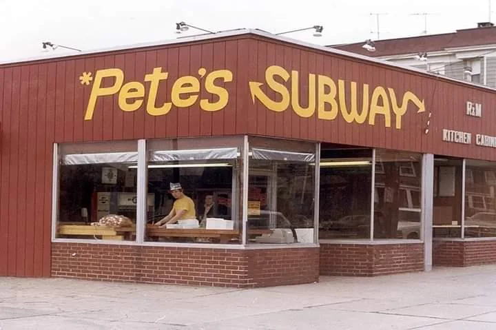 A restaurant with a red exterior and large windows displays the name "Pete's Subway" in yellow lettering. Inside, a person is working behind the counter. The street is in the foreground.