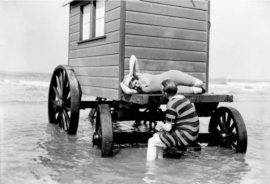 A person in an early 20th-century bathing suit lies on a wooden bathing machine above the sea. Another person in a striped swimsuit sits in the shallow water, leaning against the bathing machine's wheels. The sea and sky are visible in the background.